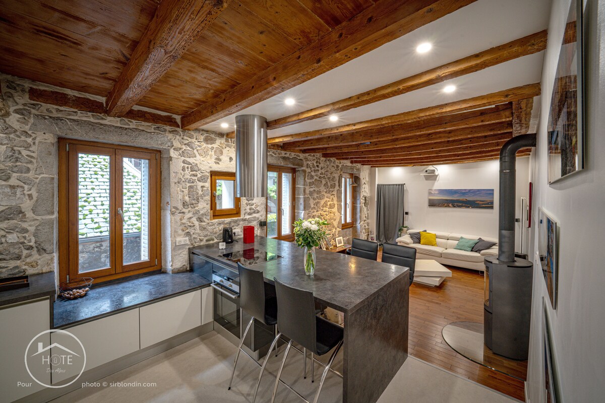 An open kitchen and living area feature exposed wooden beams and stone walls, creating a rustic ambiance. A modern kitchen island with bar seating is juxtaposed against a comfortable seating area, which includes a sofa and decorative elements. Windows provide natural light, enhancing the inviting setting.