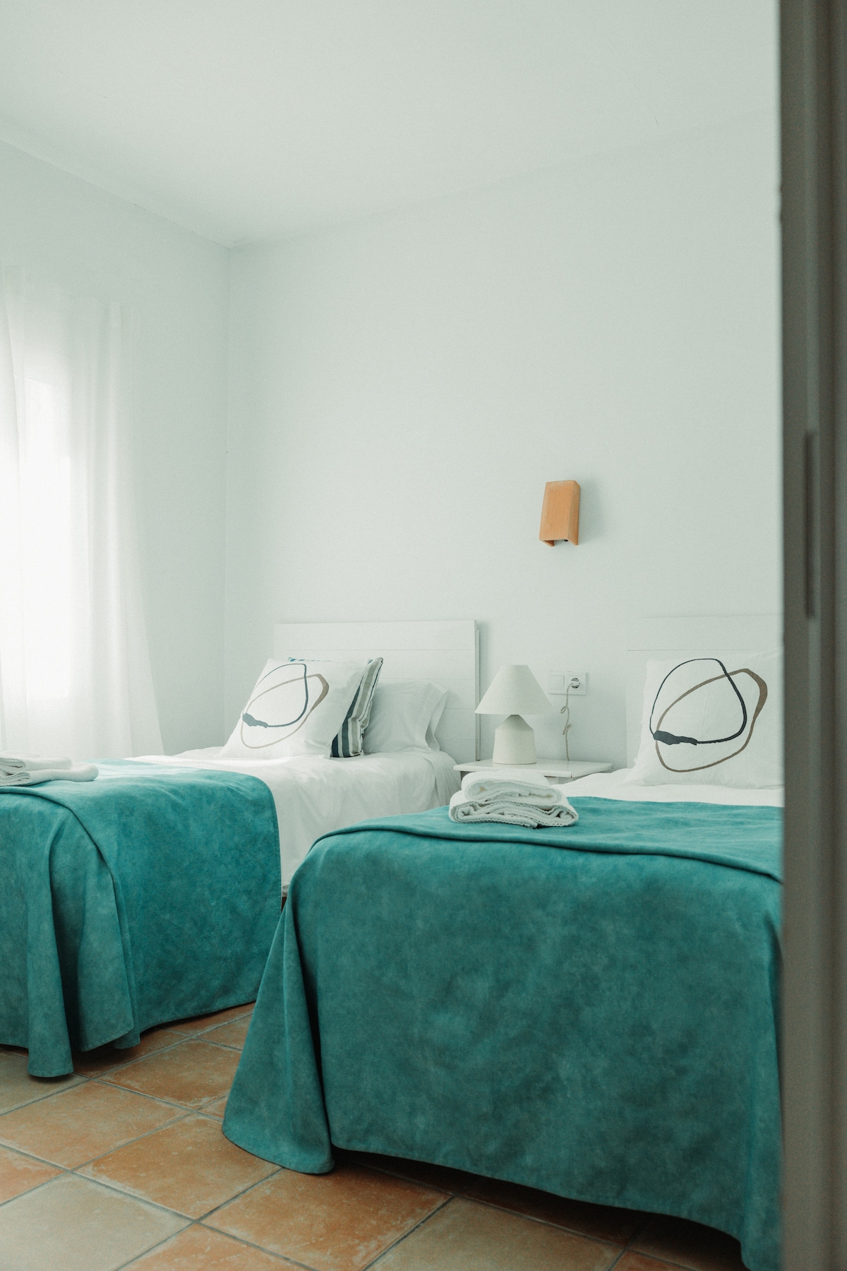 A bright bedroom features two single beds with soft teal bedspreads and decorative pillows. A small bedside lamp is placed on a white nightstand, while natural light enters through a nearby window adorned with sheer curtains.