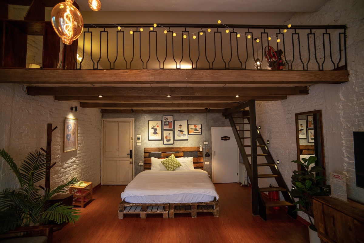 A spacious studio apartment features a comfortable sleeping area with a large bed on the lower level. A wooden loft area is visible above, accessed by a staircase. Soft lighting and artwork create a warm ambiance, while plants add a touch of greenery.