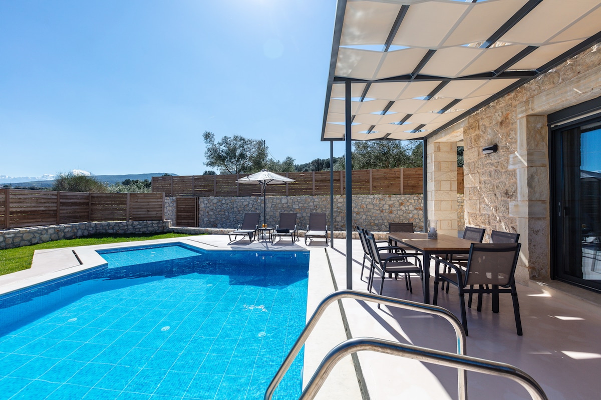 An inviting outdoor space is highlighted by a large private pool featuring clear blue water. Surrounding the pool are sun loungers under a shaded pergola. A dining area with a table and chairs is positioned nearby, offering a perfect spot for al fresco meals.