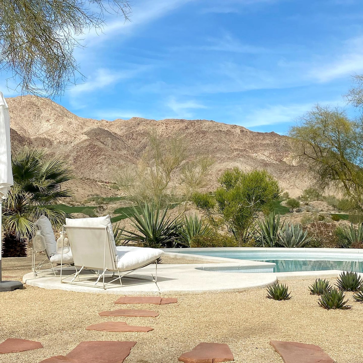 A serene patio area is shown with a white chaise lounge positioned beside a clear swimming pool. Lush desert vegetation and rocky mountains create a scenic backdrop under a clear blue sky. Pathway stones lead from the pool area to the garden.