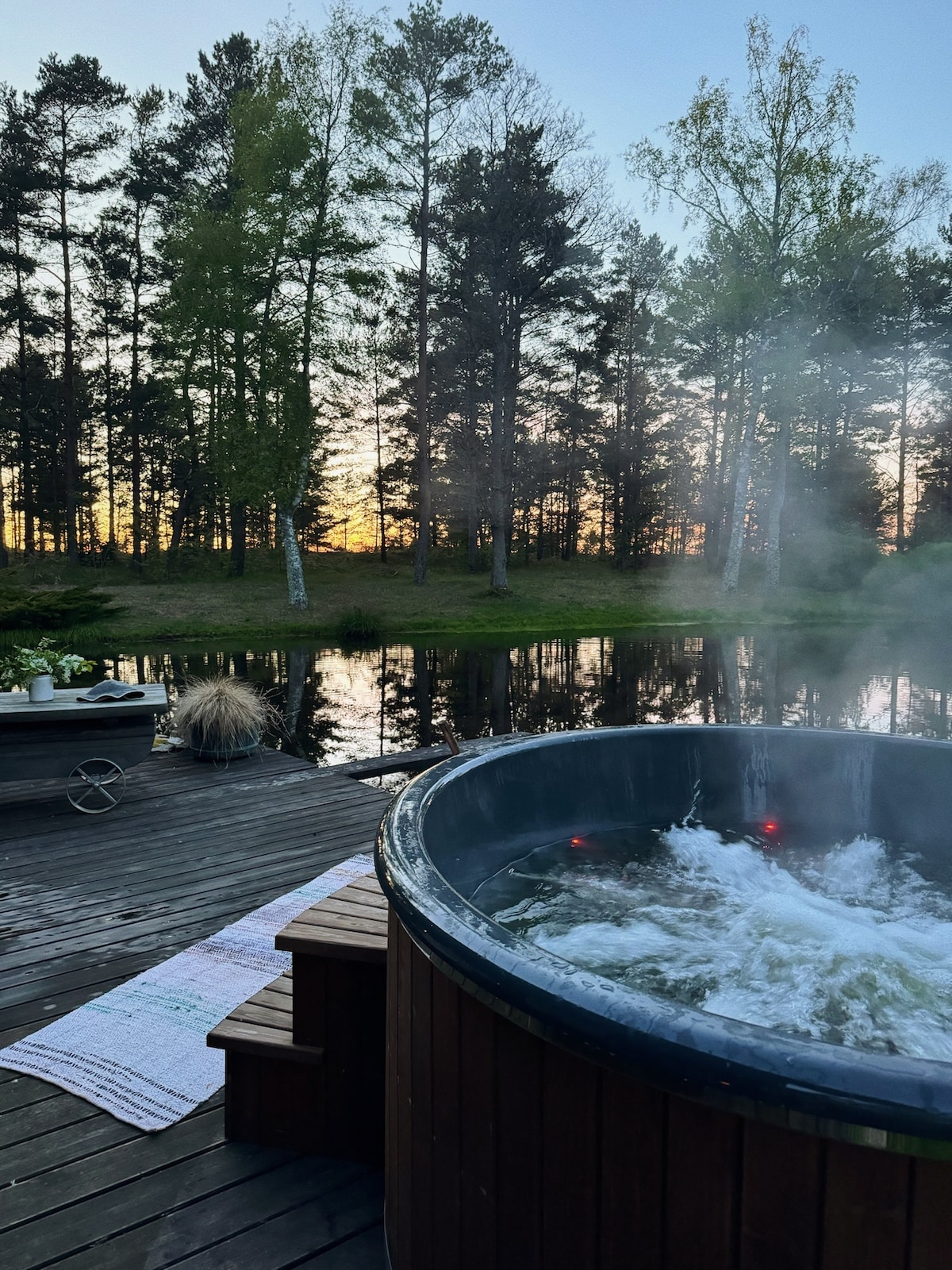 A wooden hot tub is situated on a deck overlooking a calm body of water, surrounded by lush greenery. The setting sun casts warm hues across the sky, while steam rises gently from the tub, creating a serene atmosphere.