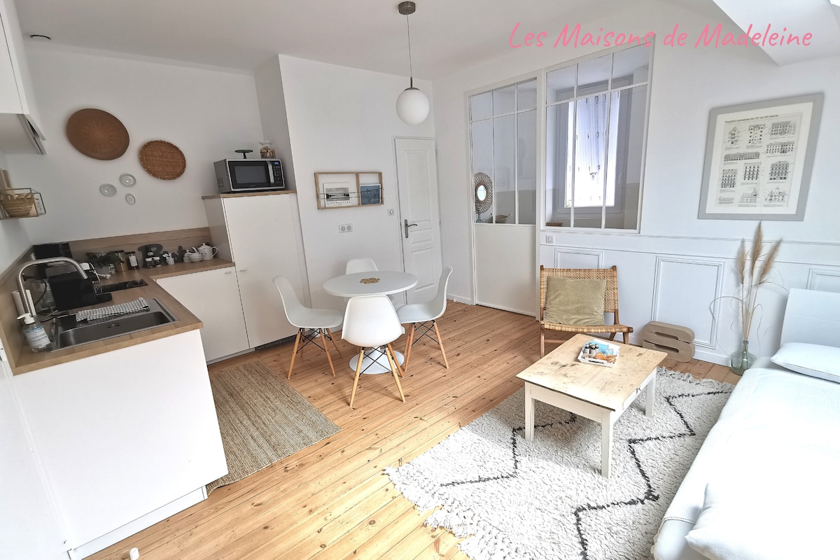 A bright and inviting living space features a compact kitchen and a dining area with a round table surrounded by white chairs. A cozy seating area includes a light-colored sofa and a simple coffee table, with natural light pouring in through the windows.