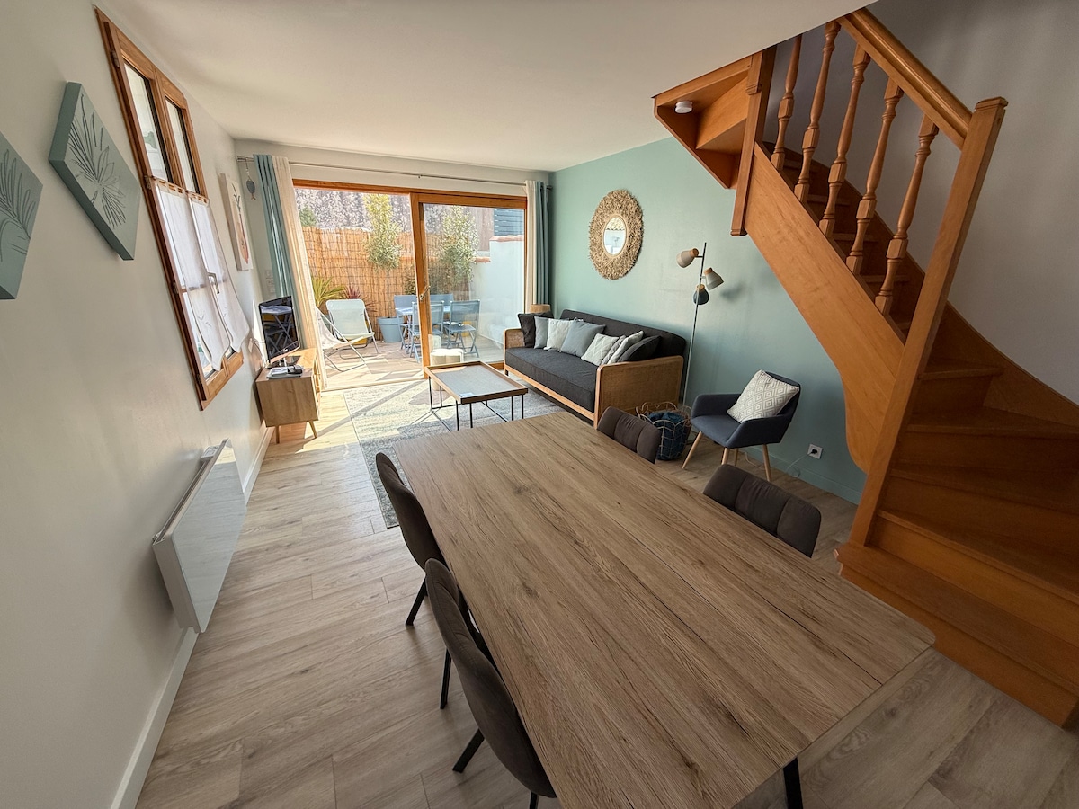A bright living area is seen, featuring a wooden dining table surrounded by chairs. A comfortable sofa and accent armchairs are positioned near large glass doors, which open up to a private outdoor space. Light wooden stairs ascend to the upper level.