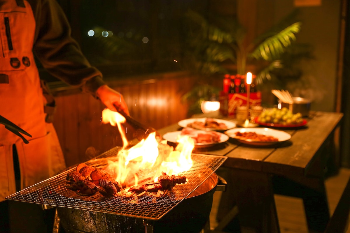 A barbecue grill is situated on a wooden table, surrounded by various dishes, including meats and vegetables. Flames are visible as food is being grilled, creating a lively cooking environment. The warm glow of the fire adds to the cozy ambiance, while foliage is seen in the background.