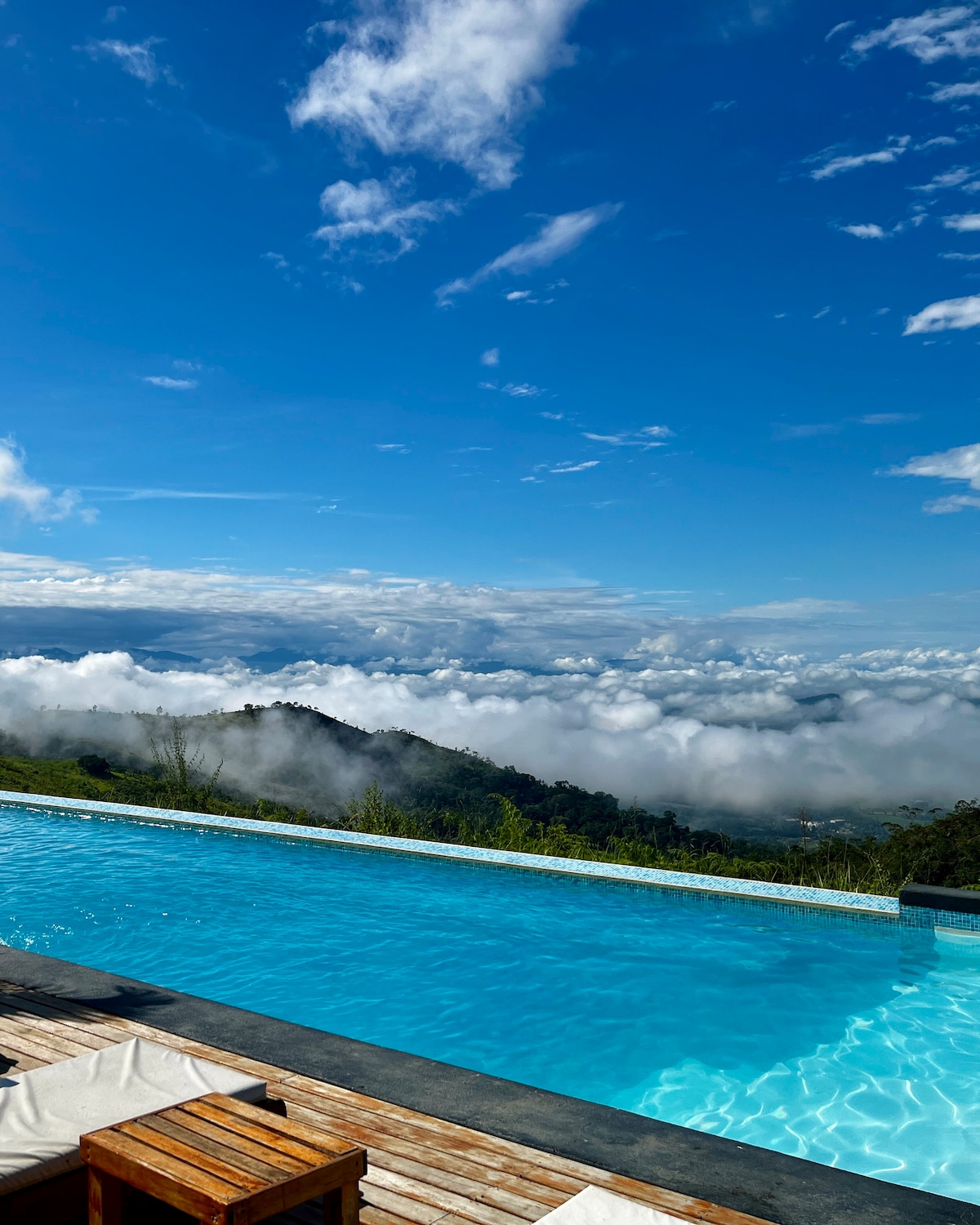 An infinity pool extends towards the horizon, offering a stunning view of clouds blanketing the lush valley below. The serene blue water reflects the clear sky, creating a seamless blend of nature and tranquility.