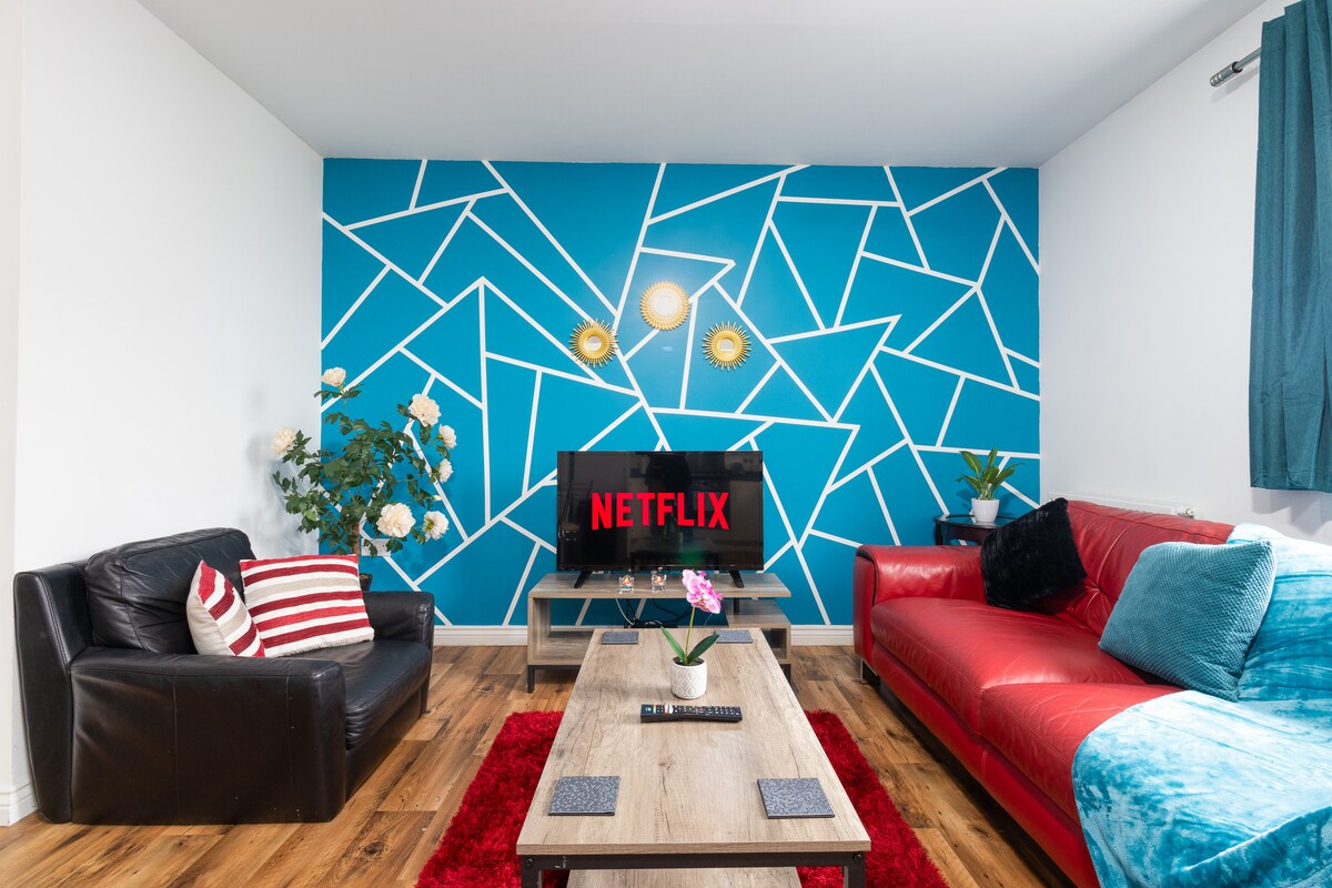 A living area features a bright blue accent wall with geometric patterns. A modern wooden coffee table sits atop a red area rug, with a black leather sofa and a vibrant red couch positioned nearby. A smart TV displaying a Netflix logo is positioned on a wooden stand.