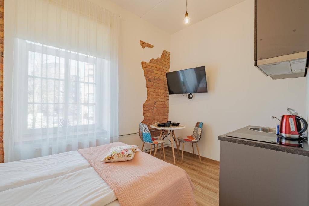 A bright room features exposed brick accent walls and a window draped with sheer white curtains. A television is mounted on the wall opposite a dining area with a small table and two colorful chairs. A kitchenette is partially visible in the corner.