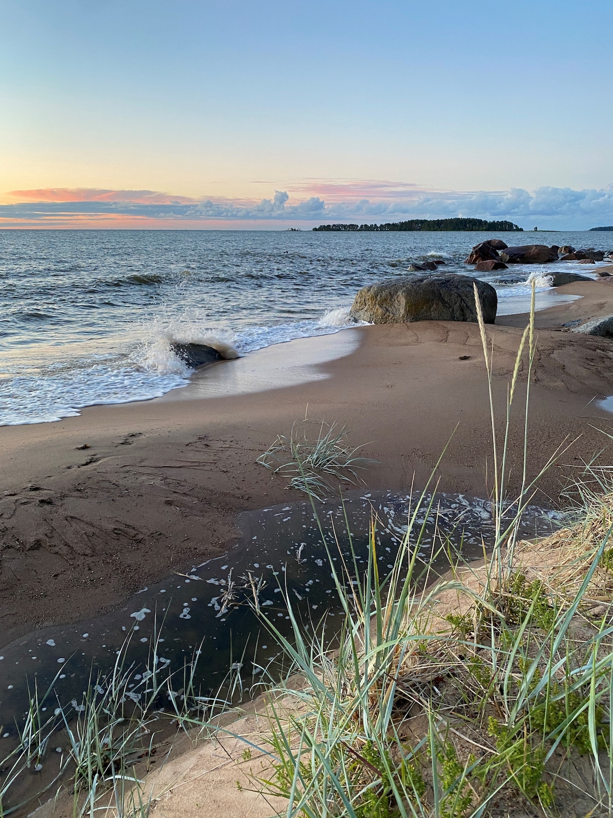 A peaceful beach scene presents gentle waves lapping against the shore, with smooth stones and sand blending harmoniously. Lush grasses line the edge, and the distant horizon displays soft hues of colors from a setting sun, complementing the calm water.