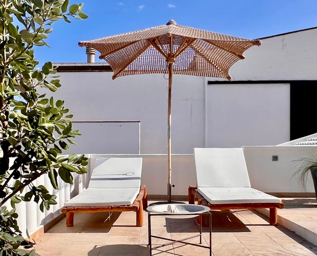 A spacious terrace is equipped with two sun loungers and a small table. A large umbrella provides shade, while surrounding greenery adds a natural touch. The area is bathed in sunlight, with a clear blue sky overhead.