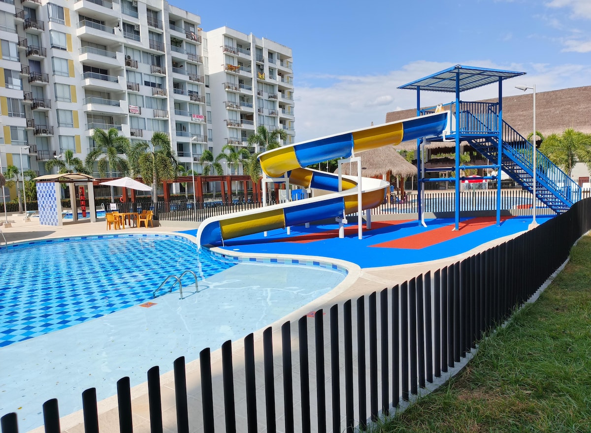 A vibrant water slide is situated adjacent to a spacious pool, creating a playful atmosphere. Colorful designs enhance the area, while lounge chairs offer seating nearby. A fence delineates the pool area, and palm trees provide greenery in the background under a clear blue sky.