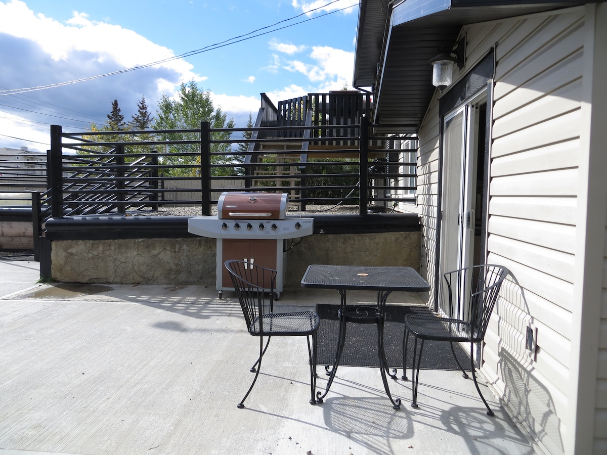An outdoor patio area features a small table with two black metal chairs, providing a space for relaxation. A grill stands nearby on a stone surface, and a wooden railing offers a sense of separation from the surrounding environment. Trees are visible in the background.