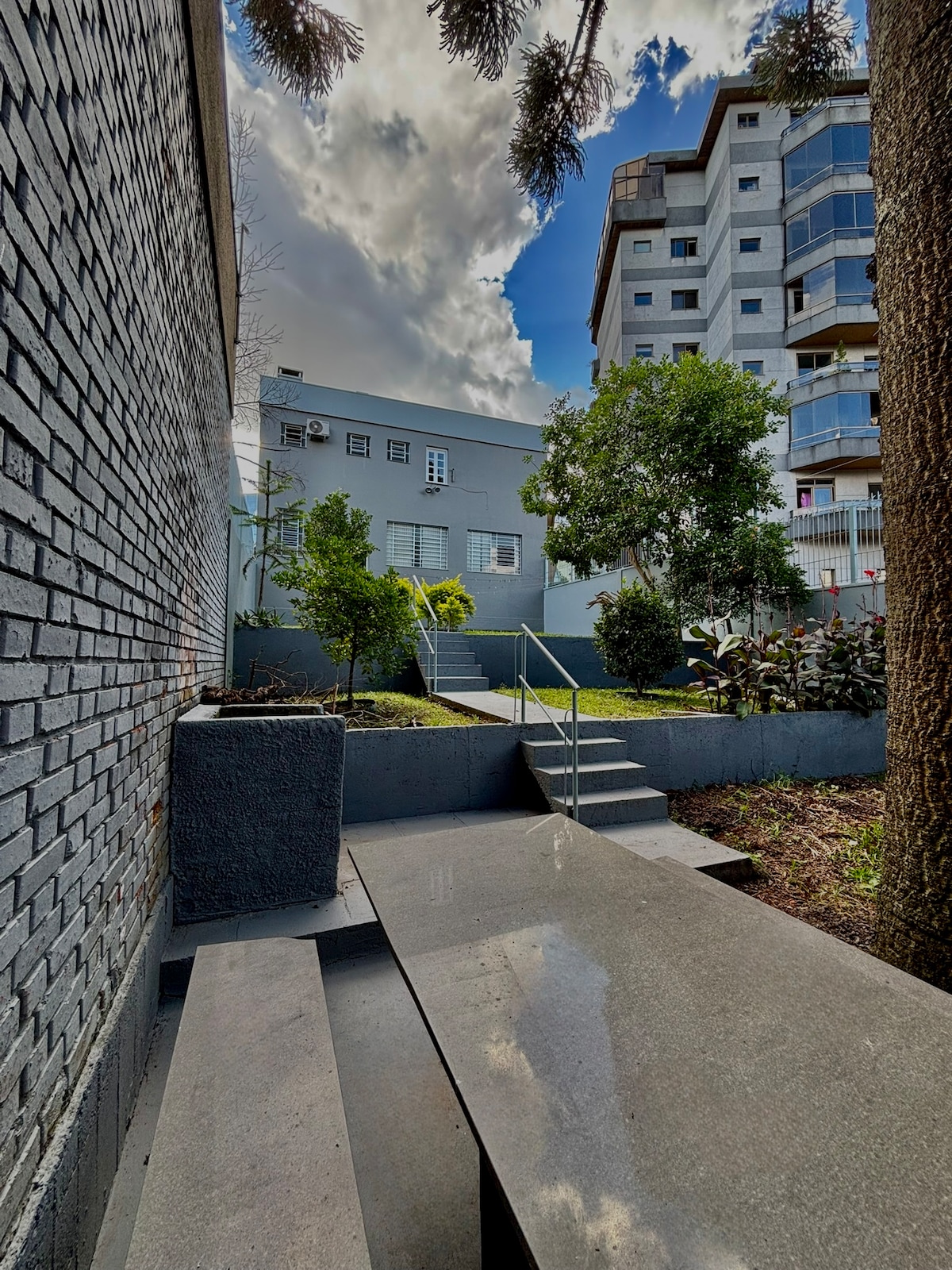 A garden area is shown with a grey stone pathway leading to a set of steps. Surrounding greenery includes trees and shrubs, enhancing the tranquil setting. The sky is partly cloudy, creating a serene atmosphere over the well-maintained outdoor space.