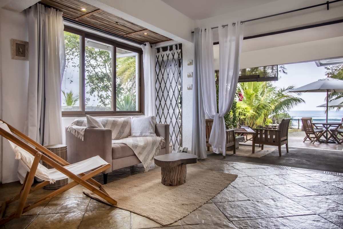 A welcoming living area features a comfortable sofa draped with a soft throw, accompanied by a rustic wooden coffee table. Large windows allow natural light to enter, offering views of the beach and surrounding greenery. Sheer curtains gently frame the space, enhancing its openness.