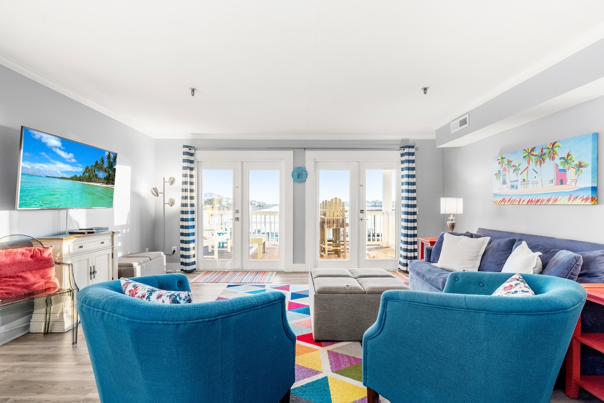 A light-filled living area showcases large glass doors that open to water views. Two comfortable blue chairs are positioned around a multi-colored area rug. A soft gray ottoman sits at the center, while a TV is mounted on the wall, and vibrant artwork adorns the space.