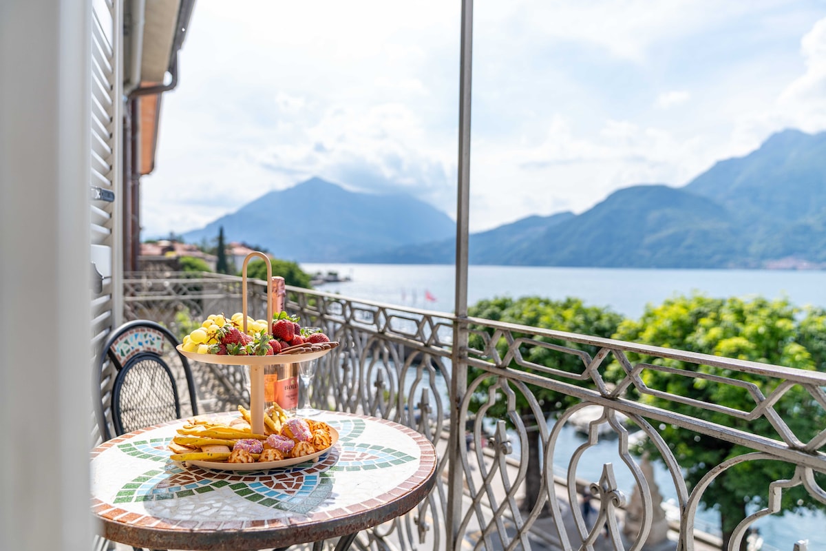 A terrace table holds a tiered fruit platter featuring fresh grapes, strawberries, and assorted pastries. The backdrop includes scenic views of the lake and surrounding mountains, with soft clouds and greenery framing the tranquil atmosphere.