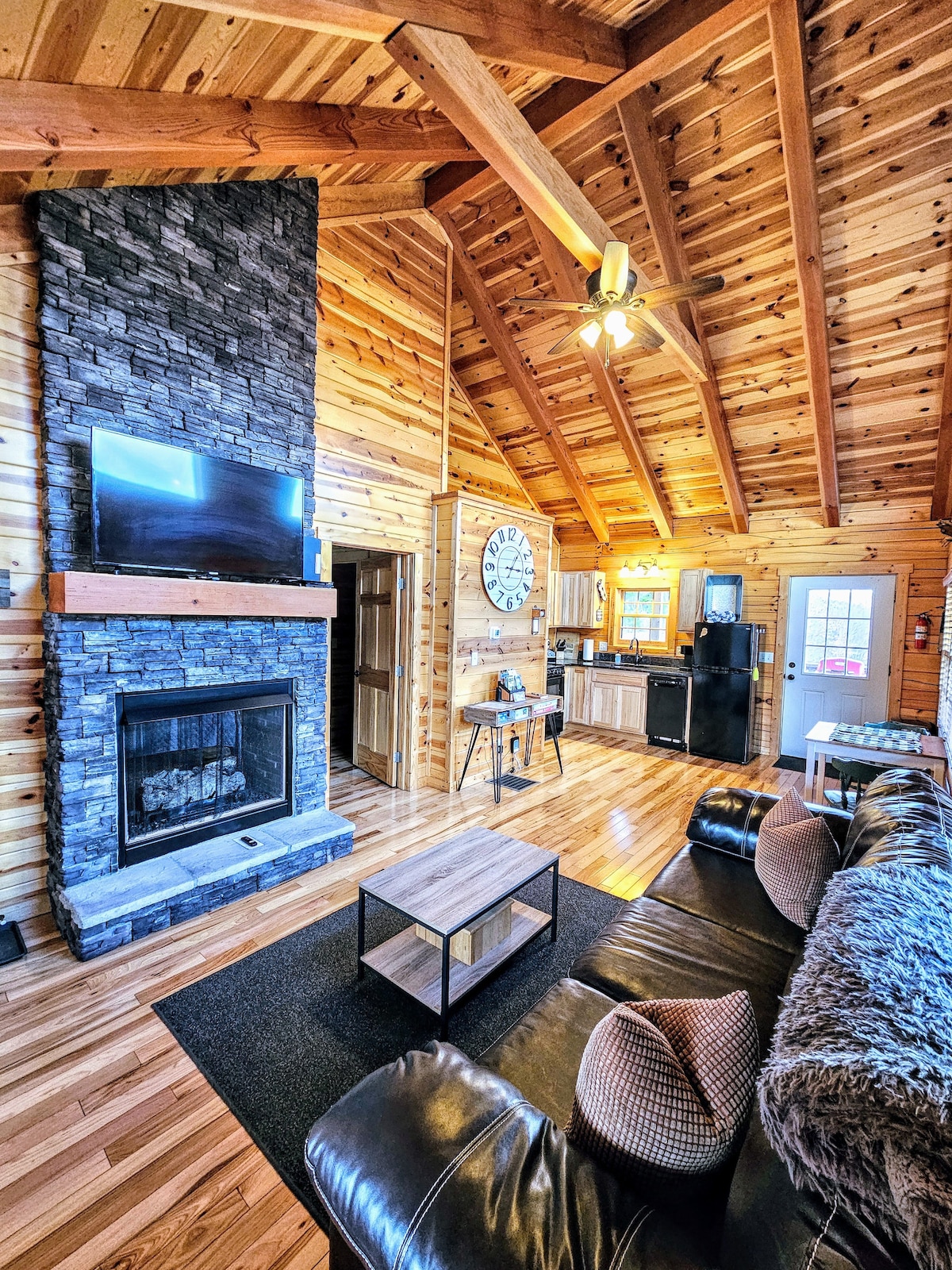 An open living area is featured with vaulted wooden ceilings and a stone fireplace. A television is mounted above the fireplace. A sofa with decorative pillows is positioned in front of a contemporary coffee table. Light streams in from adjacent windows, illuminating the wooden surfaces.