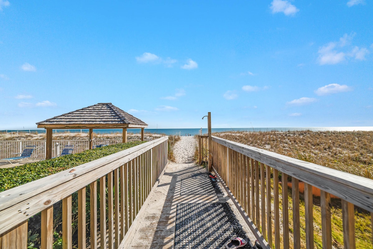 A wooden walkway leads to a serene beach, framed by vegetation and a gazebo. Ocean waves can be seen in the distance under a bright blue sky, creating an inviting pathway for guests to enjoy private beach access.