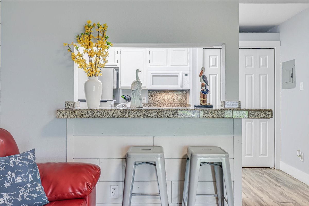 A kitchen area is visible through an open passthrough, featuring a granite bar with two metal stools. A vase with yellow flowers adds a touch of color. The kitchen is equipped with white cabinetry and appliances, providing a clean and functional look.