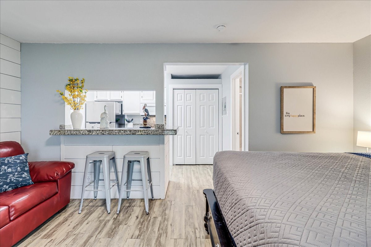 The room features a queen-sized bed with a textured blanket, accompanied by a red sofa. A small kitchen bar with three stools is visible, set against a neutral color scheme. Light flows in from an unseen window, enhancing the space's cozy layout.