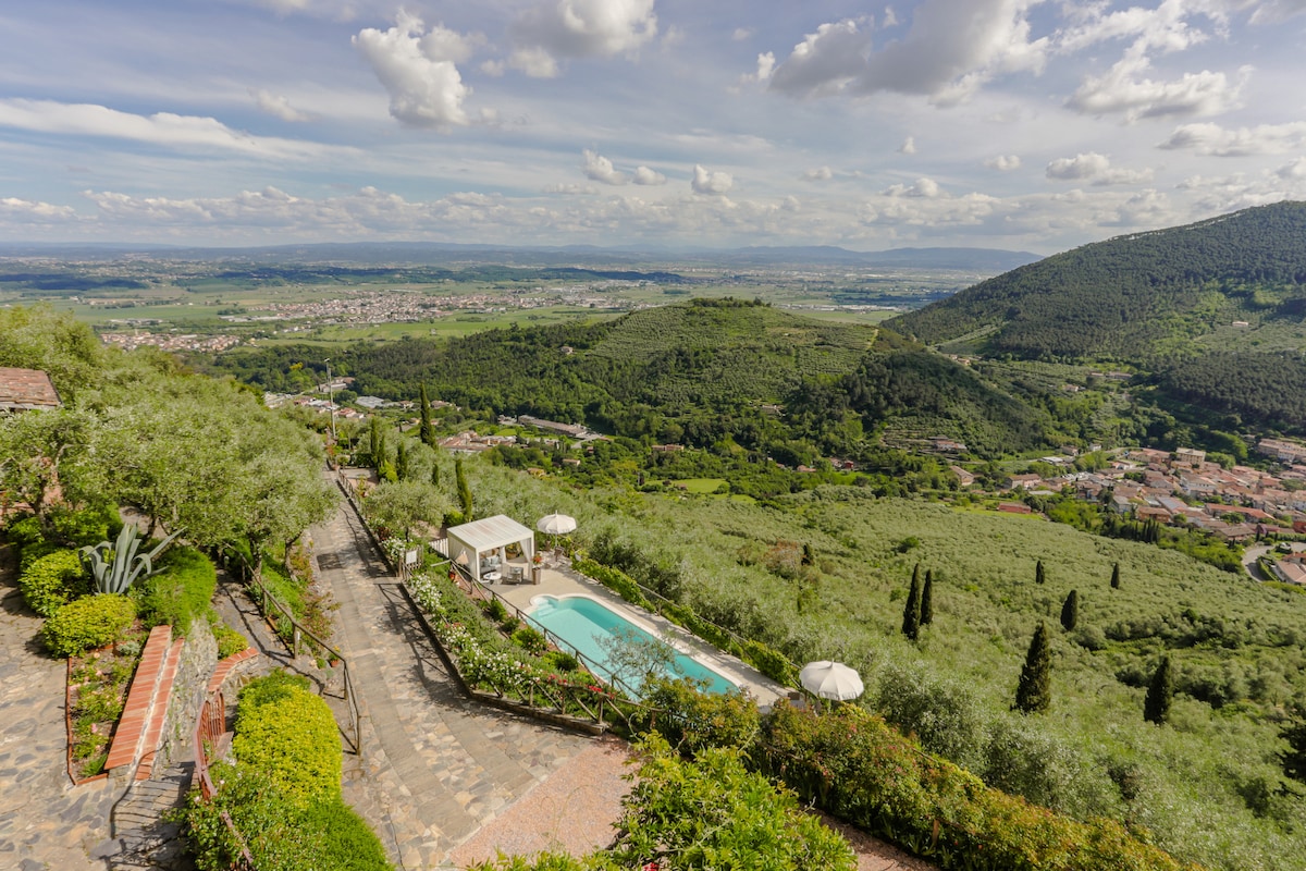 A panoramic view captures rolling hills adorned with olive groves, overlooking a tranquil pool area. A shaded gazebo rests beside the pool, while cypress trees gently rise in the background. The landscape is rich with greenery, offering a serene escape amidst nature.