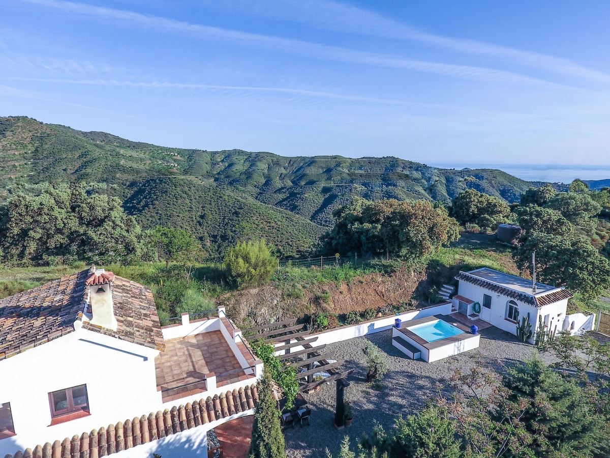 The image captures the expansive landscape surrounding the finca, showcasing rolling green hills and trees under a clear blue sky. The property features a private plunge pool, sun terraces, and a white structure integrated harmoniously into nature, with outdoor seating areas visible nearby.