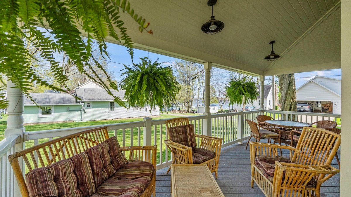 A covered porch features comfortable seating with a striped sofa and multiple woven chairs. Hanging ferns add greenery, while a wooden table invites outdoor dining. The view includes a spacious lawn and neighboring homes, contributing to a relaxed atmosphere.