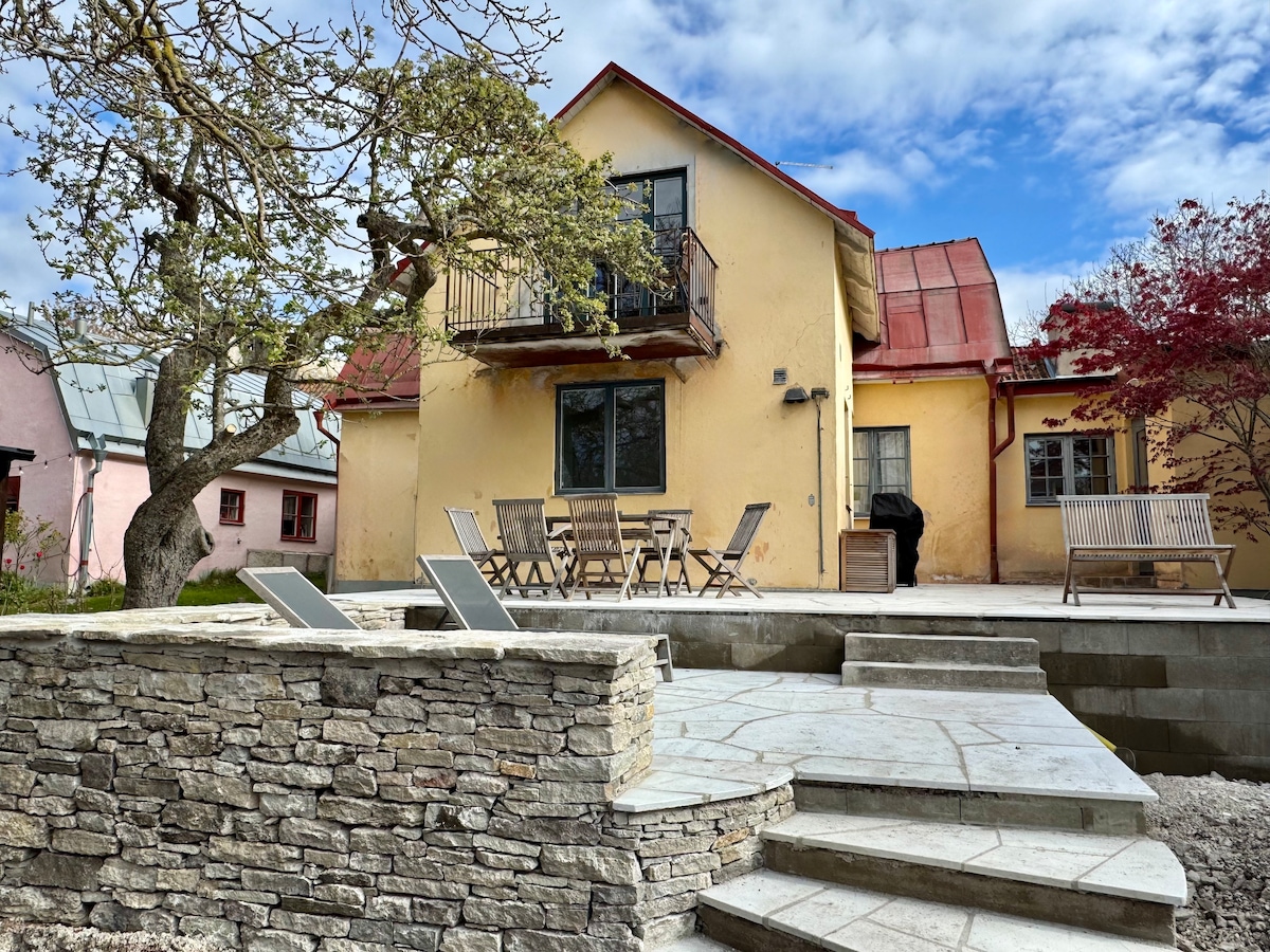 A charming yellow house with a red roof is viewed from a stone patio. Outdoor seating, including a dining table and lounge chairs, enhance the space. A tree provides shade, and a grill is positioned nearby, completing the outdoor setting.