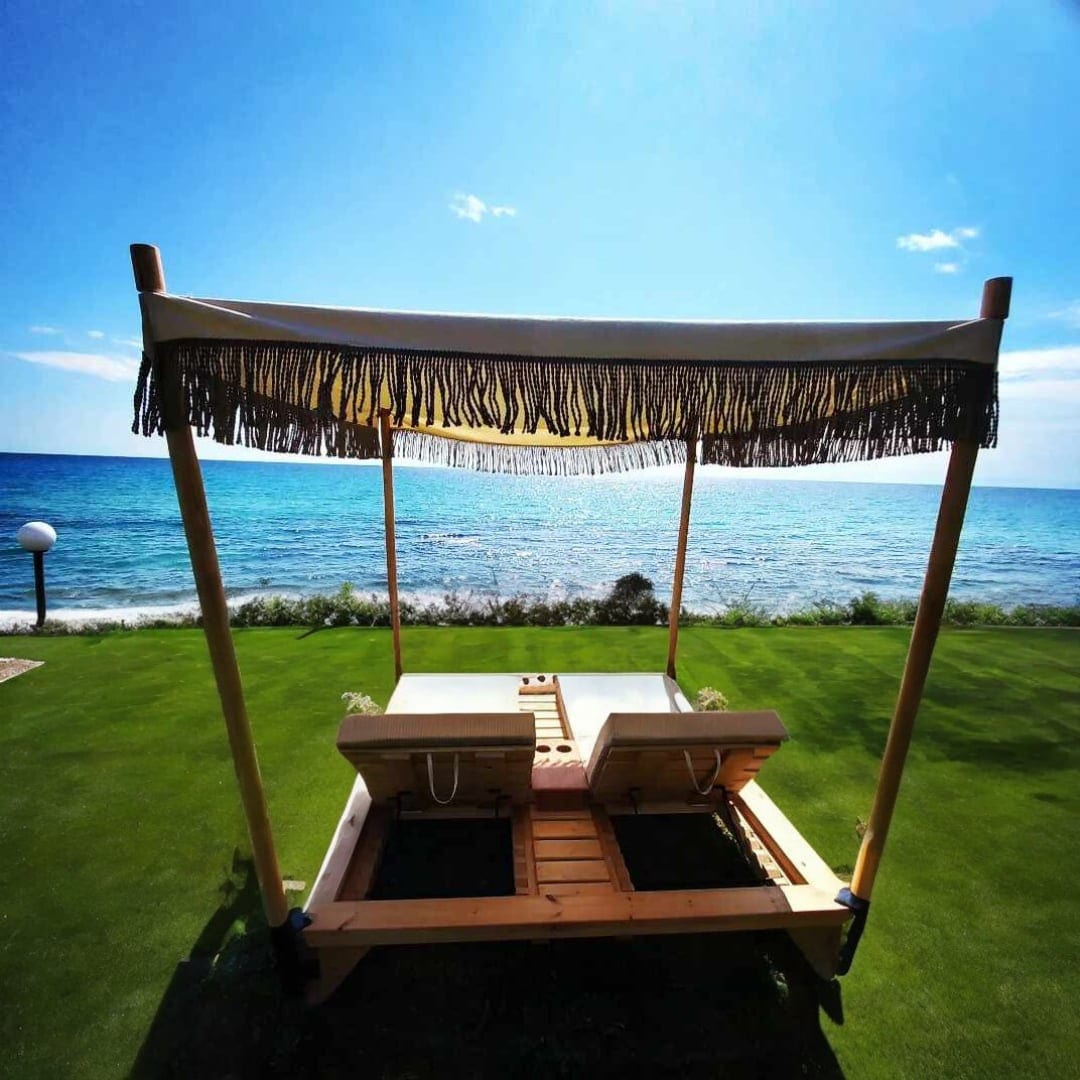 A shaded wooden lounge area featuring two comfortable sunbeds sits on a lush green lawn, facing the serene sea. The vibrant blue sky complements the tranquil waters, creating a calming space for relaxation.