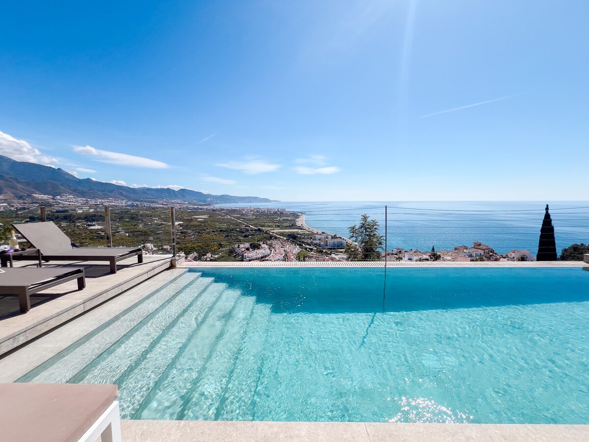 An infinity pool extends towards a panoramic view of the sea and coastline, framed by the mountains in the background. Sun loungers are positioned near the edge of the pool, inviting relaxation in the serene atmosphere created by the clear blue sky.