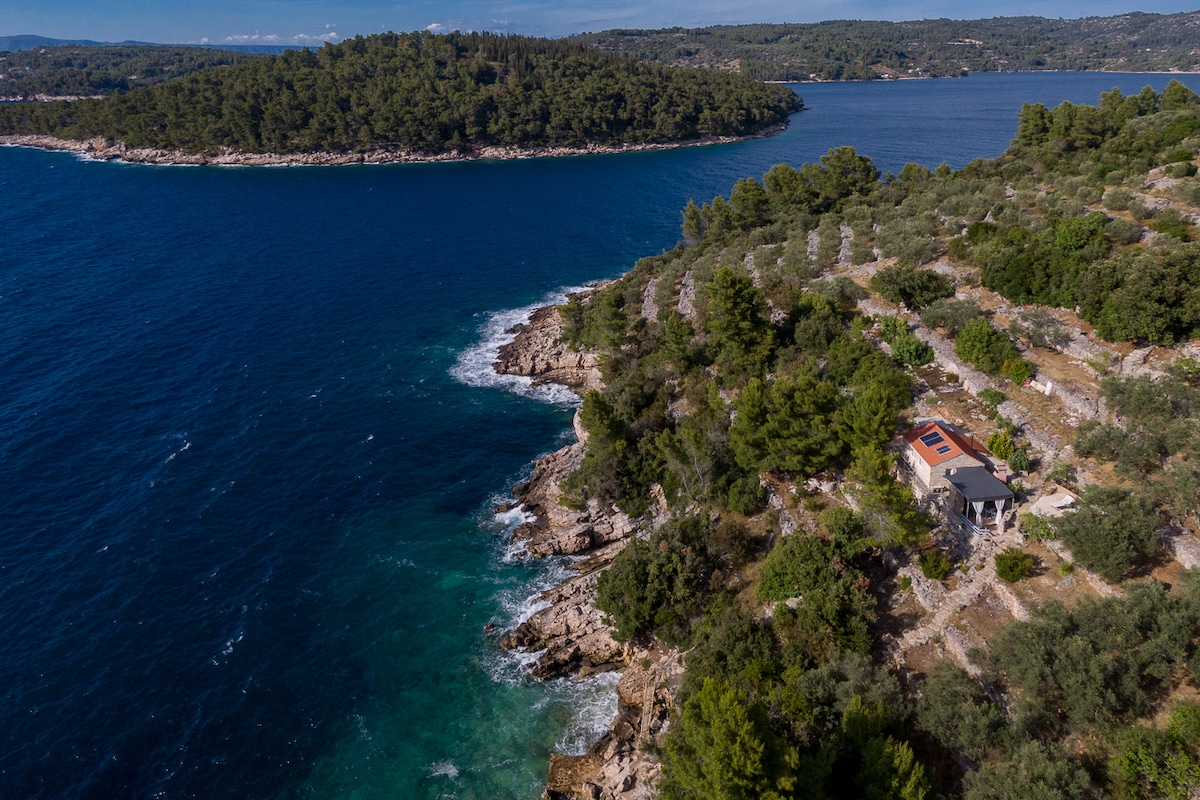 An aerial view showcases a small stone house situated on a rocky coastline. Surrounding natural greenery and olive trees provide a serene backdrop, while the turquoise waters gently lap against the shore, highlighting the tranquil cove.