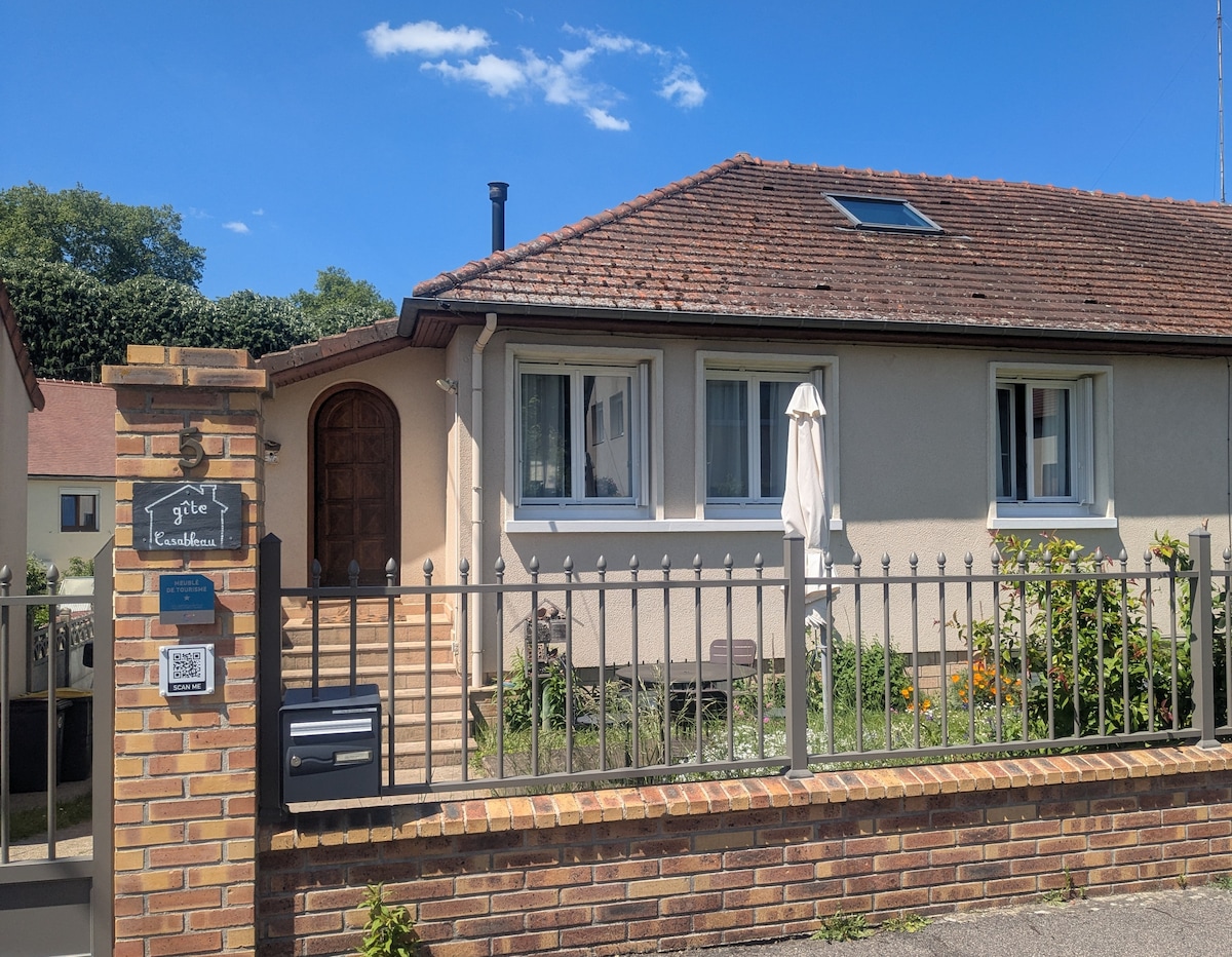 The exterior of the gîte features a charming single-story building with a red-tiled roof and a welcoming entrance. A gated fence surrounds the property, and a patio umbrella is visible in the garden area. A sunny blue sky complements the green landscaping.