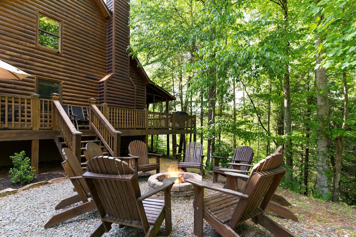 An outdoor fire pit surrounded by wooden Adirondack chairs is positioned amidst a forested setting. The log cabin stands nearby, featuring a spacious deck that offers a connection to nature. Lush greenery provides a serene backdrop to the gathering area.
