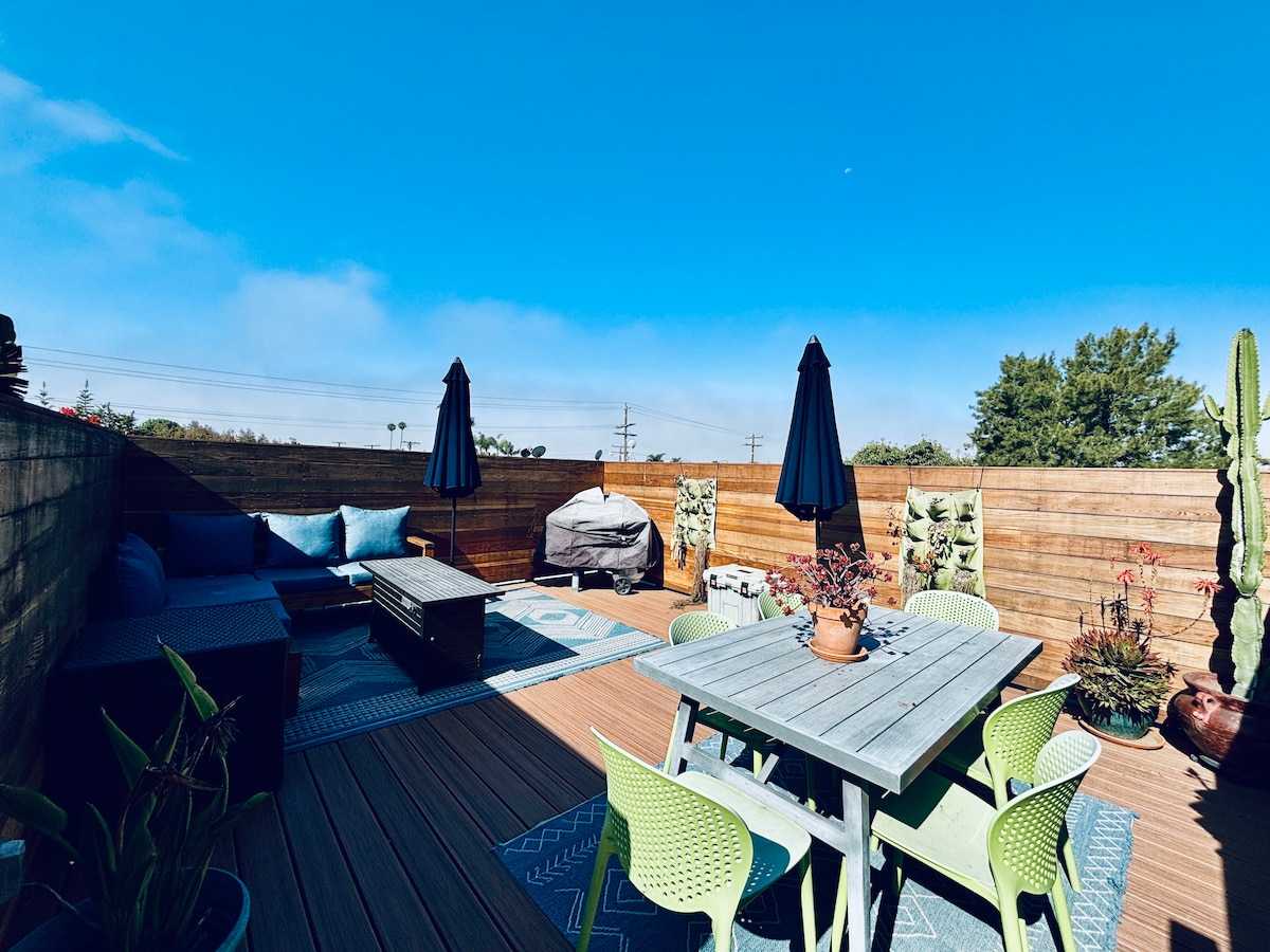 A rooftop deck is showcased, featuring a dining table surrounded by light green chairs. A lounge area with cushions is present, while two large umbrellas provide shade. The space is bordered by wooden panels and adorned with potted plants, against a clear blue sky.
