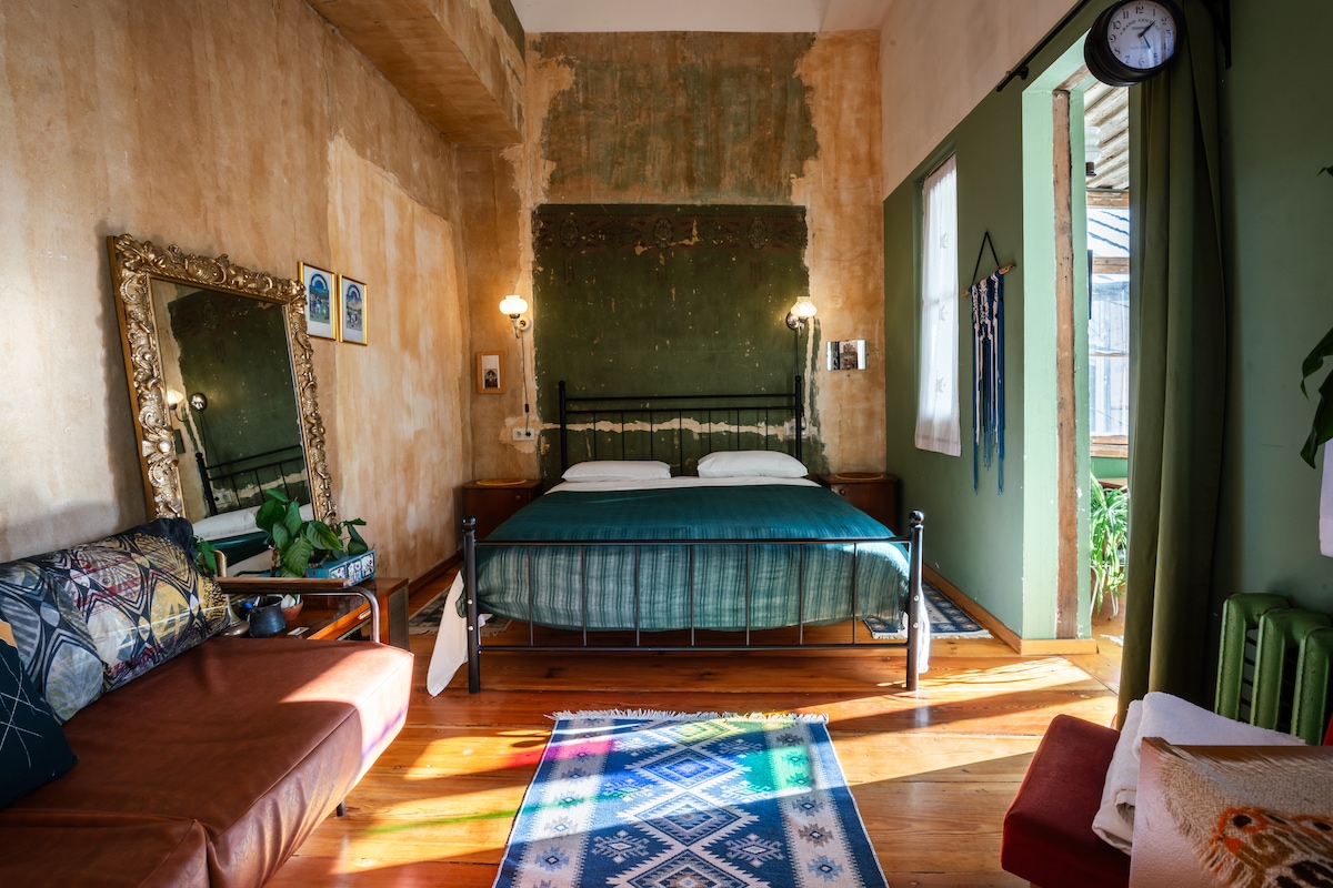 A bright and spacious room is furnished with a black metal bed featuring a green bedspread. A cozy couch is positioned to one side, with a colorful rug on the wooden floor. Natural light enters through the window, highlighting the textured walls and decorative mirror.