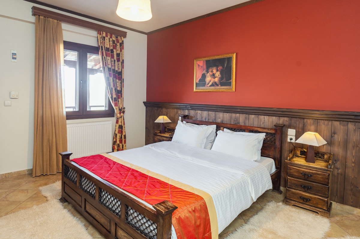 A spacious bedroom is presented with a large bed featuring a red and white throw blanket. Wooden nightstands are positioned on either side, each with a lamp. A window with stylish curtains allows natural light to enter, and a warm-toned wall enhances the overall ambiance.