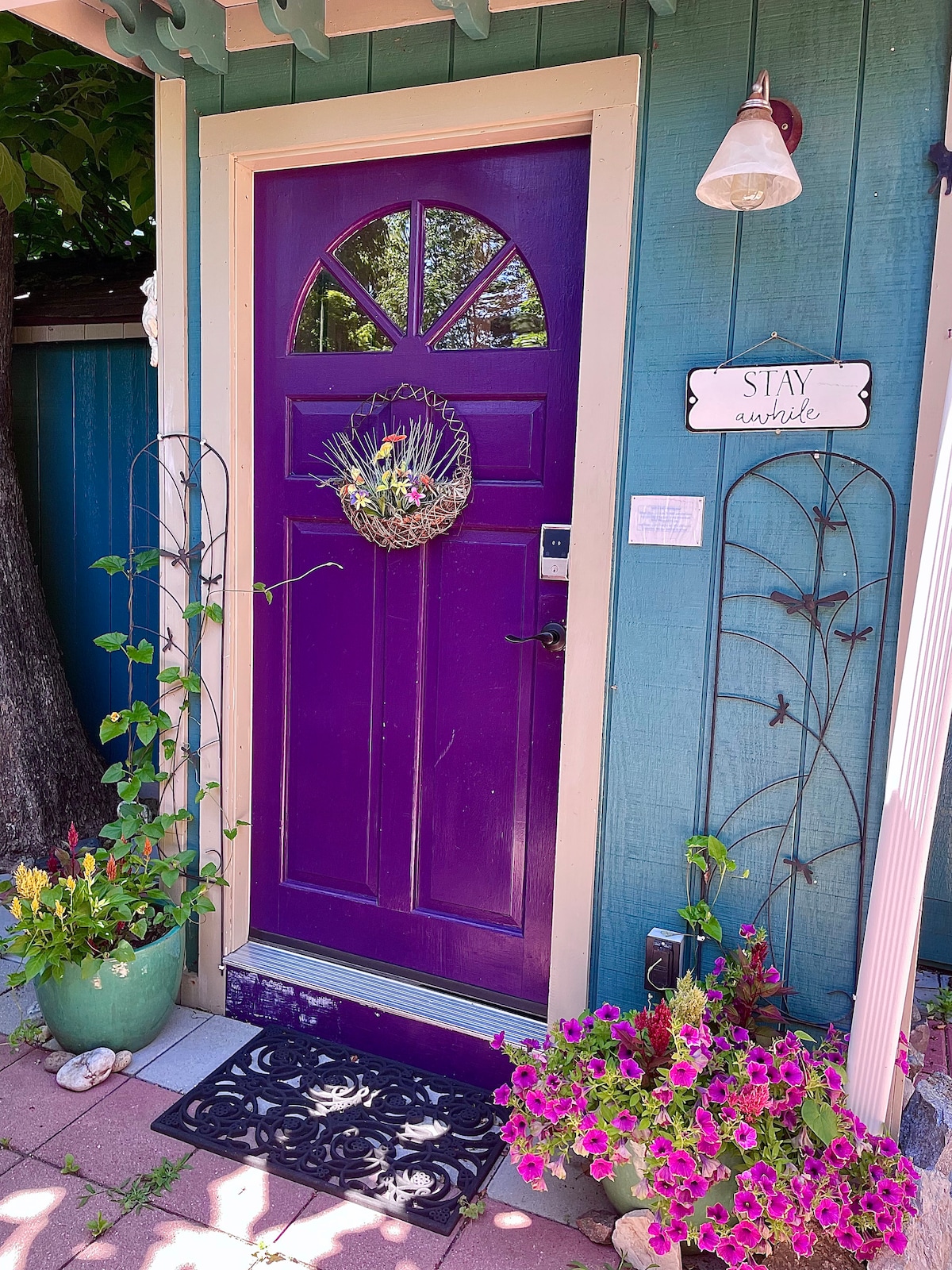 A vibrant purple door welcomes guests, adorned with a decorative wreath and accompanied by two potted plants. A black welcome mat is placed at the entrance. The surrounding walls are painted in a soothing aqua color, complemented by a nearby light fixture and decorative wall sign.