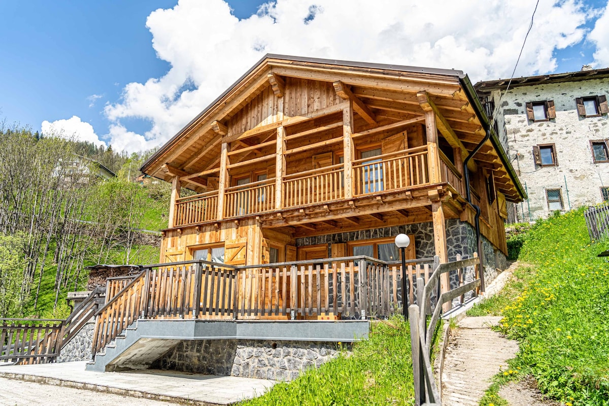 The chalet features a rustic wooden façade with a spacious balcony and multiple large windows that invite natural light. A stone pathway leads to the entrance, flanked by lush green grass and wildflowers, providing an inviting outdoor area for relaxation.
