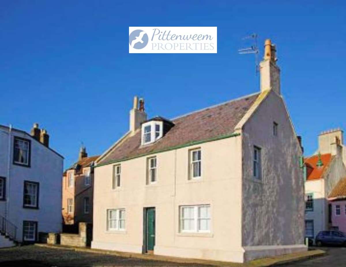 The exterior of a charming two-story house is presented, featuring a soft pink façade under a clear blue sky. Windows are framed in white, while the roof displays a combination of tiles and a chimney. Nearby buildings create a quaint village atmosphere.