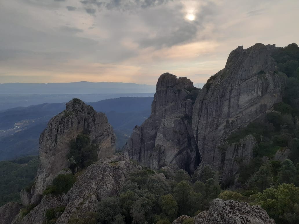 Rugged rock formations rise prominently against a soft, dusky sky. The landscape features a mix of dense greenery at the base, with distant mountains visible in the background. The subtle interplay of light creates a serene atmosphere, emphasizing the natural beauty of the area.