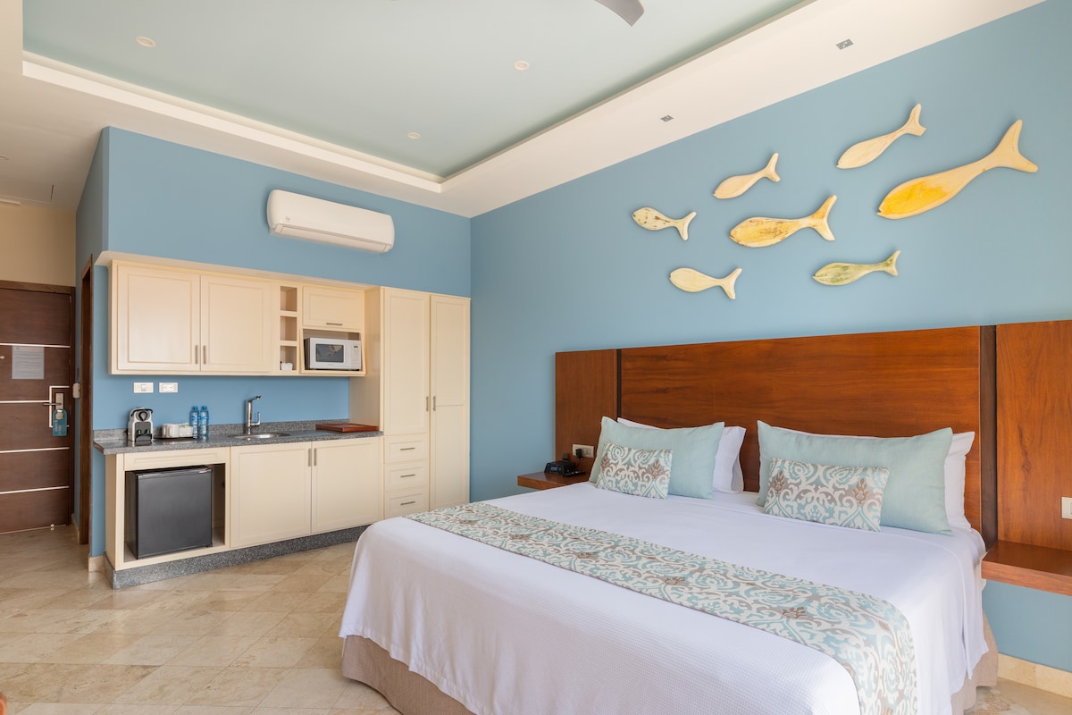 A spacious guest room features a large bed with light linens and decorative pillows. A kitchenette is visible, equipped with a microwave and refrigerator. Colorful fish wall art adds a decorative touch, while a cool blue wall creates a calm atmosphere. Natural light fills the space from windows.