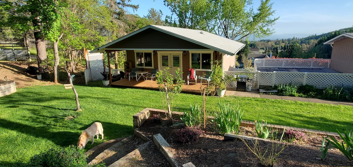 The exterior of the unit is shown, featuring a private deck with seating and a view of the surrounding greenery. A well-maintained lawn is visible, along with flower beds and mature trees, which enhance the tranquil country setting.