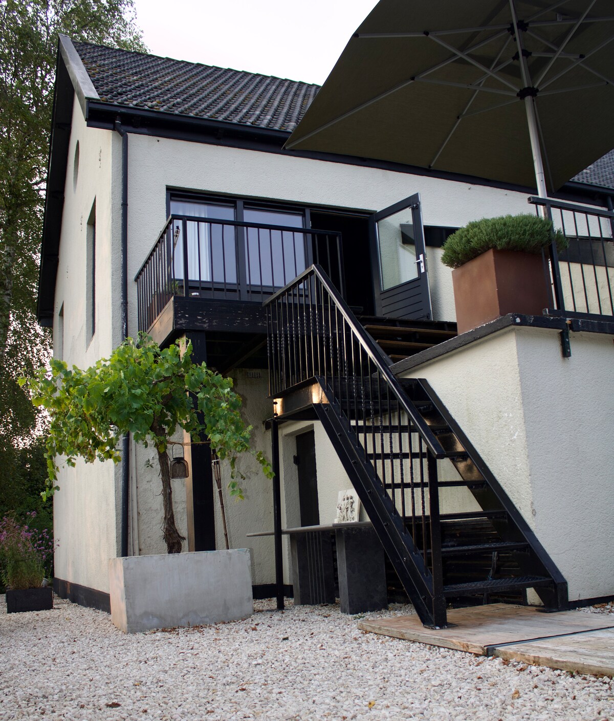 The exterior of a detached house is presented, featuring a staircase leading to a balcony. Black railings and potted greenery enhance the entryway, while a large umbrella provides shade. A gravel pathway surrounds the house, contributing to its inviting outdoor space.