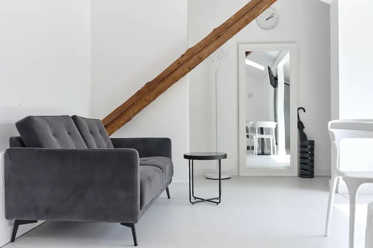 A cozy living area is furnished with a gray plush sofa positioned near a slender black side table. A tall mirror reflects the bright space, complemented by a wooden beam that adds warmth. A minimalist coat rack and umbrella stand are also present.