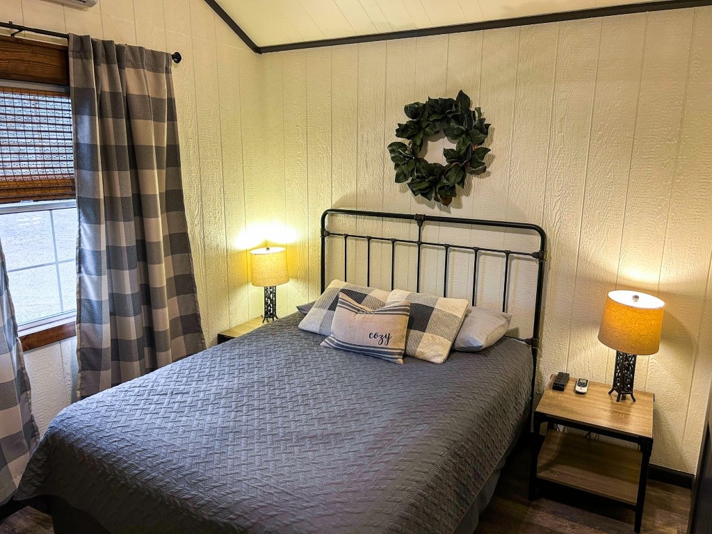 A cozy sleeping area features a metal frame bed topped with a quilted grey coverlet. Two bedside lamps provide soft illumination, and decorative pillows add contrast. A wreath hangs on the wall, complementing the warm tones of the curtains and the natural light from the window.