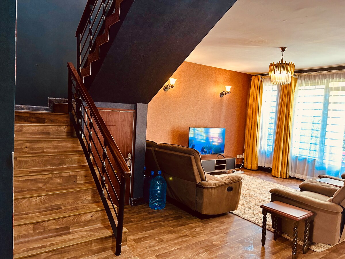 The living area features a comfortable seating arrangement with light-colored upholstery, positioned near a television. Natural light enters through large windows, complemented by soft yellow curtains. A staircase leads to the upper level, and a small wooden table is positioned beside the seating.