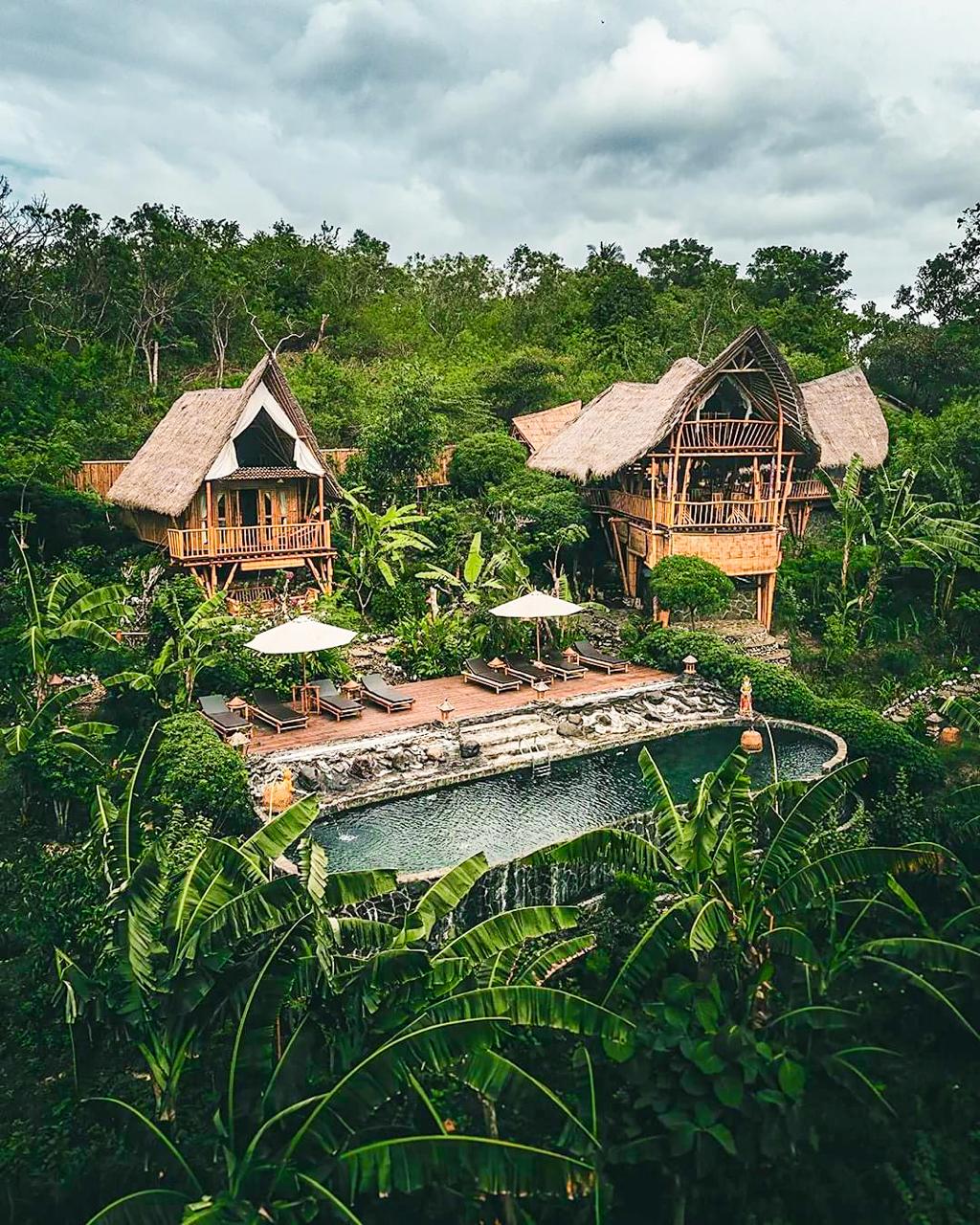 The image depicts two bamboo resort structures nestled among lush greenery, with thatched roofs allowing for natural ventilation. A swimming pool is bordered by a wooden deck featuring sun loungers and umbrellas, surrounded by vibrant tropical plants, creating a serene ambiance.