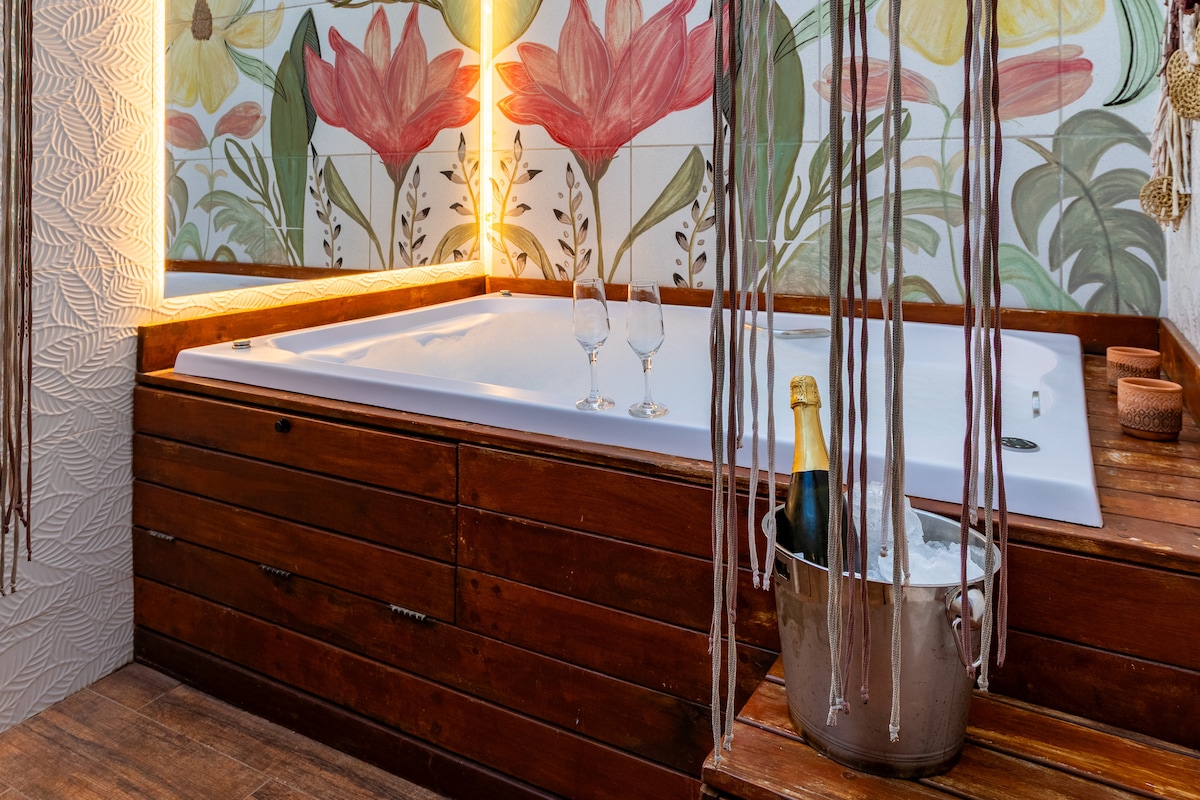 A warm and inviting jacuzzi is set against a vibrant floral mural, creating a peaceful spa-like retreat. Nearby, a bucket filled with champagne rests on the wooden platform, complemented by two elegant champagne glasses, ready for a relaxing experience.