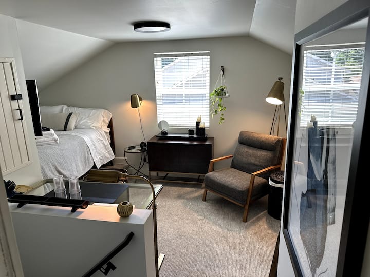 Dormitorio de concepto abierto con TV, cama doble, armario empotrado y cajones, escritorio, silla de estar, cargadores universales de teléfono, cristalería y un reloj Alexa. 