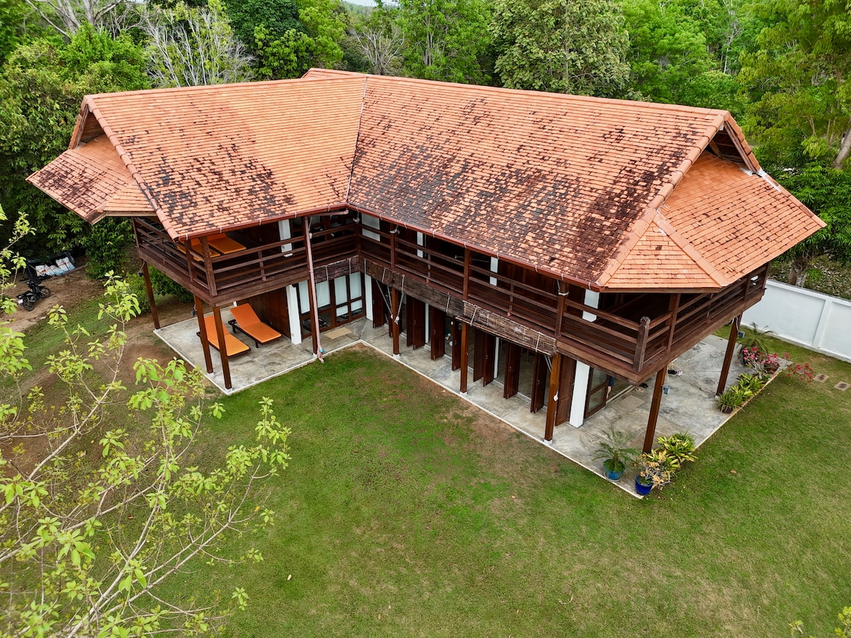 The villa's exterior is showcased from an aerial view, highlighting the traditional wooden architecture and terracotta roof tiles. Surrounding the structure, a well-maintained lawn and lush greenery create a serene environment. Several outdoor lounge areas, including two orange loungers, are visible on the patio.